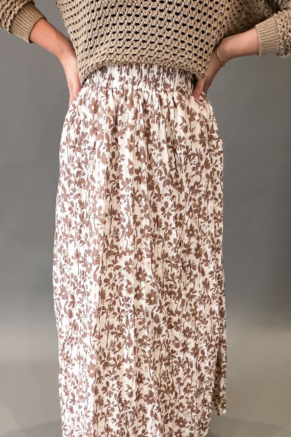 A person wearing a long pleated skirt with a brown floral pattern on a cream background, paired with a beige knit top, shown from the chest down against a neutral gray studio background.