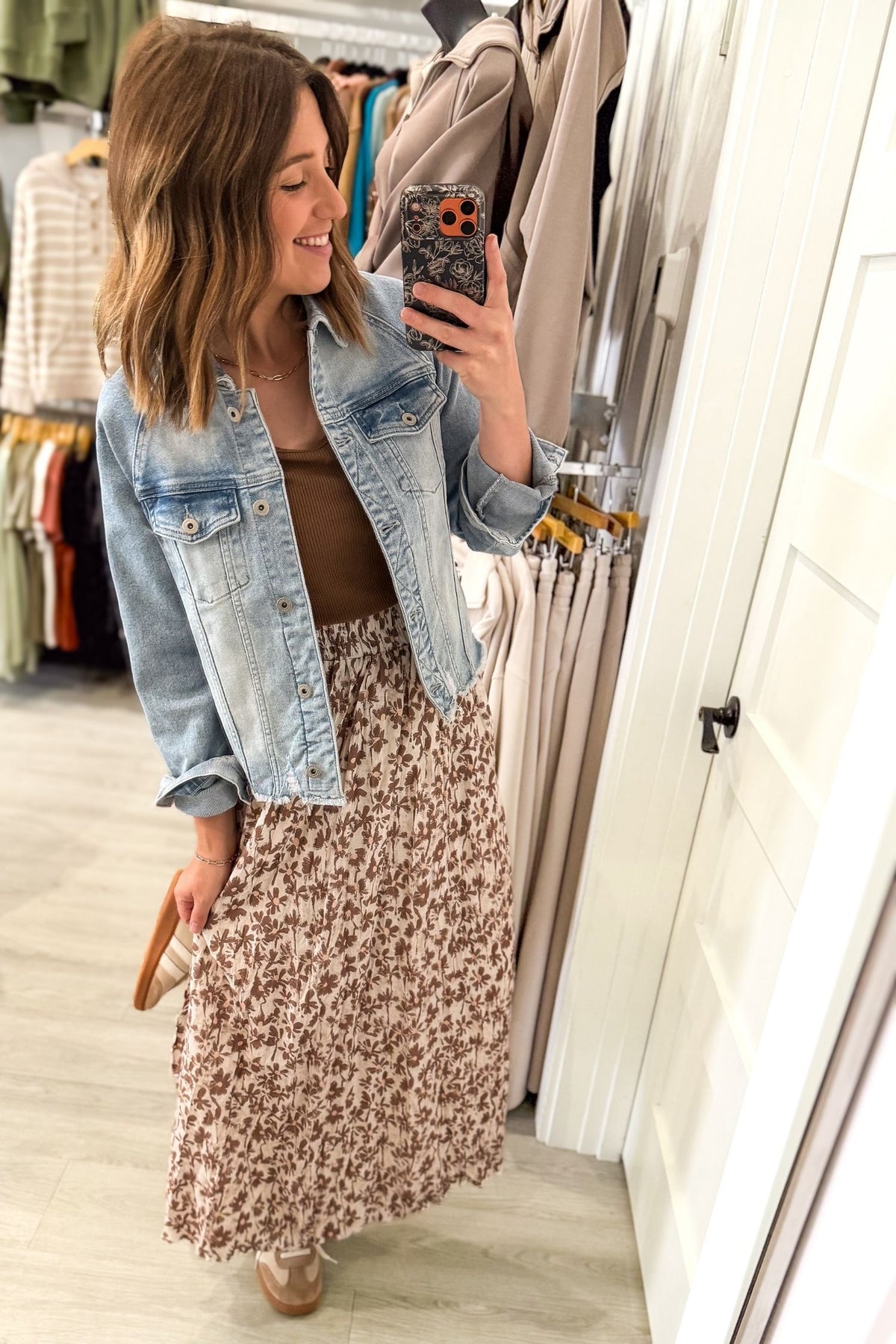 A woman takes a mirror selfie in a clothing boutique, wearing a light-wash blue denim jacket over a brown top and a brown and beige floral print maxi skirt.