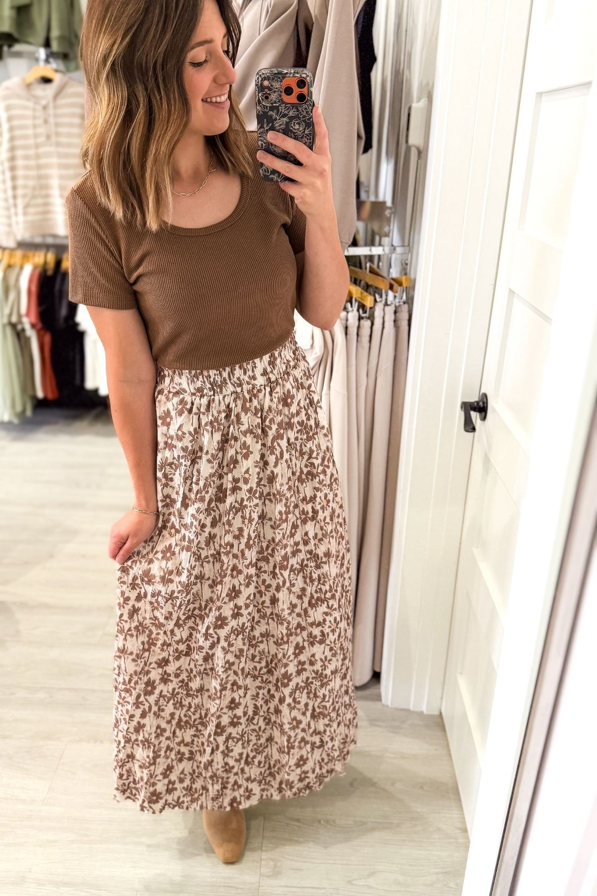 A woman takes a mirror selfie in a clothing store, wearing a brown short-sleeve scoop-neck top and a high-waisted, knee-length maxi skirt with a brown and beige floral pattern.