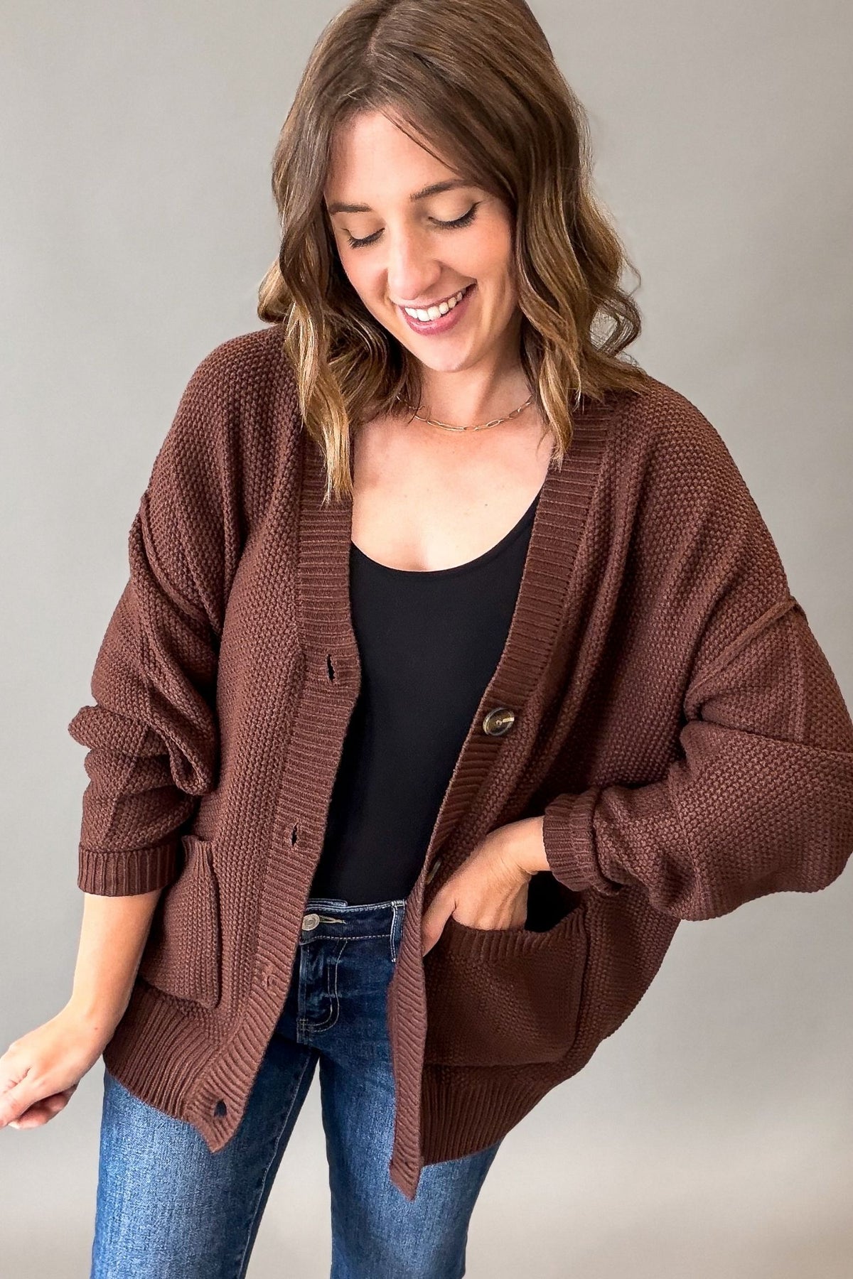 A woman wearing a brown waffle-knit cardigan over a black top and blue jeans, standing against a neutral gray background.