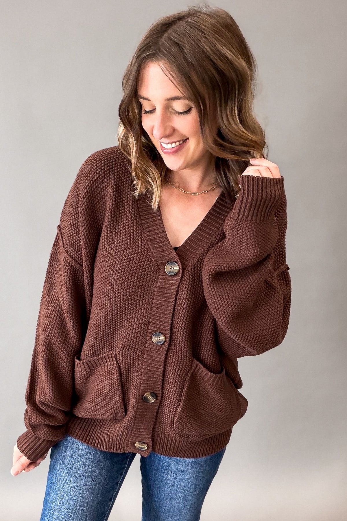 A woman wearing a brown waffle-knit cardigan sweater with front buttons and large patch pockets, paired with blue jeans, photographed against a plain gray studio background.