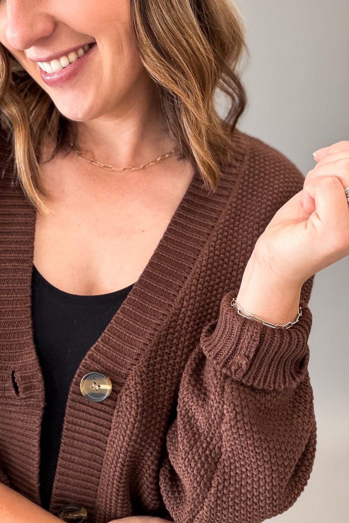 A close-up of a woman wearing a brown textured knit cardigan over a black top, with a neutral gray background.