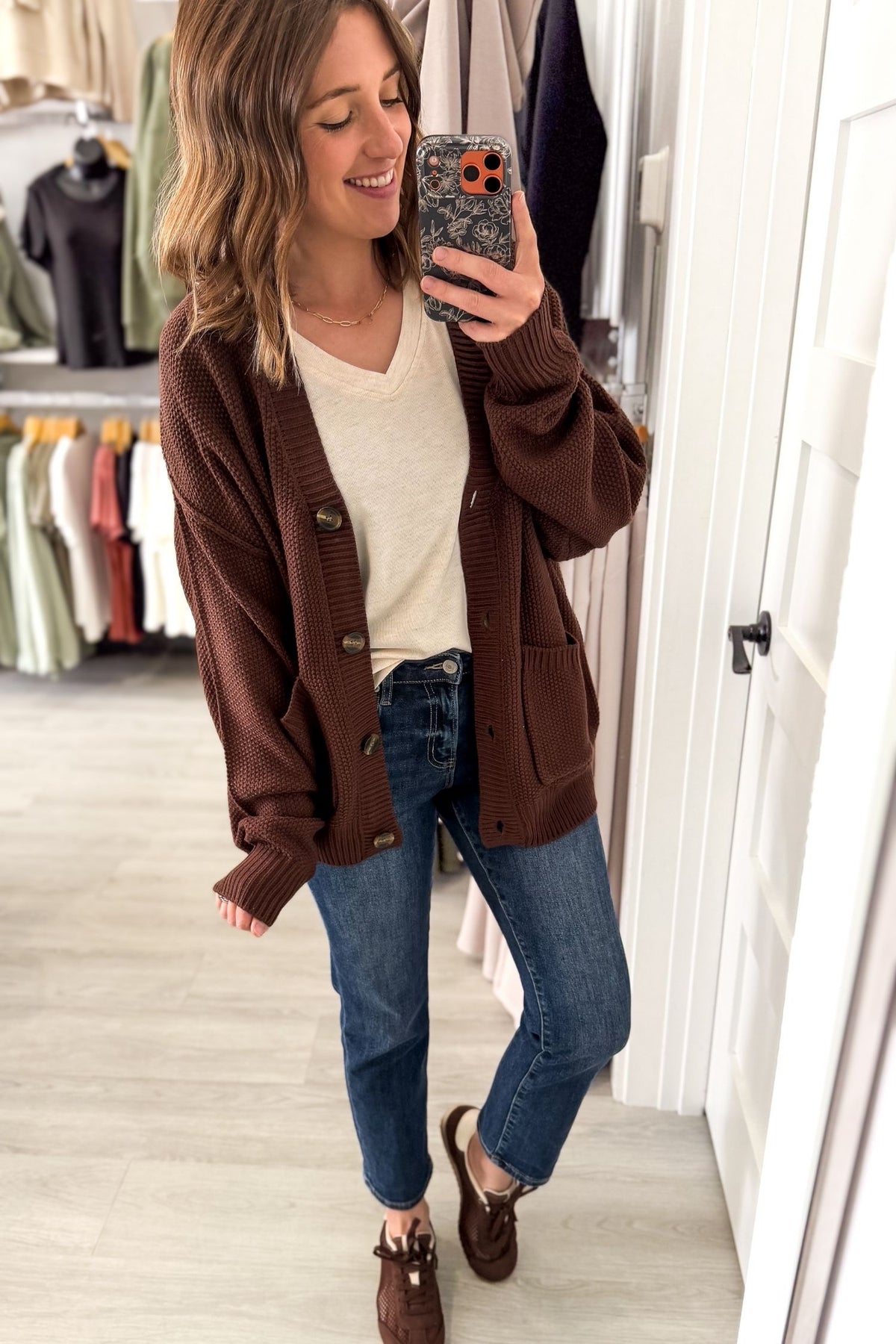 A woman takes a mirror selfie in a boutique, wearing a brown ribbed knit cardigan over a cream V-neck top, blue jeans, and brown loafers.