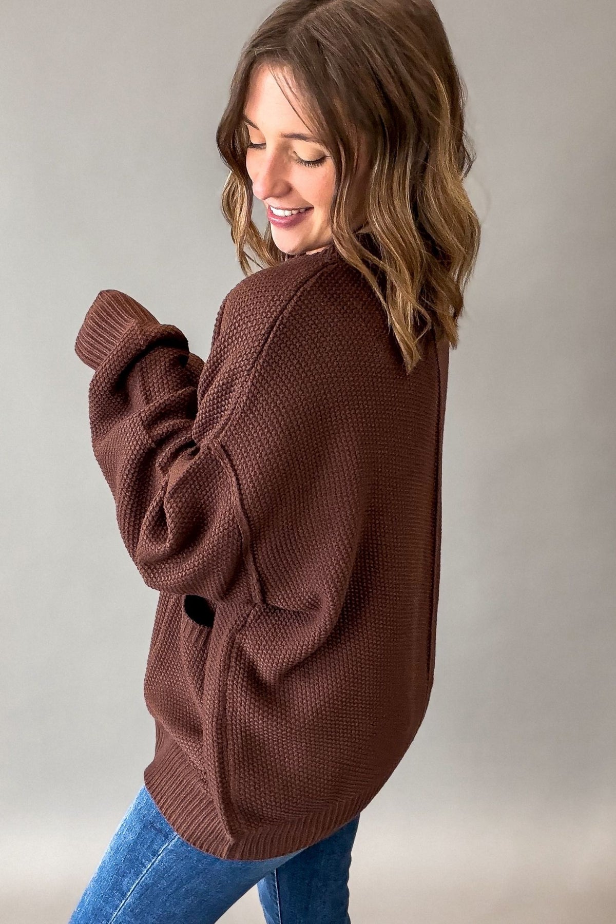 A woman wearing a brown waffle-knit sweater and blue jeans, photographed from the side against a plain light gray studio background.
