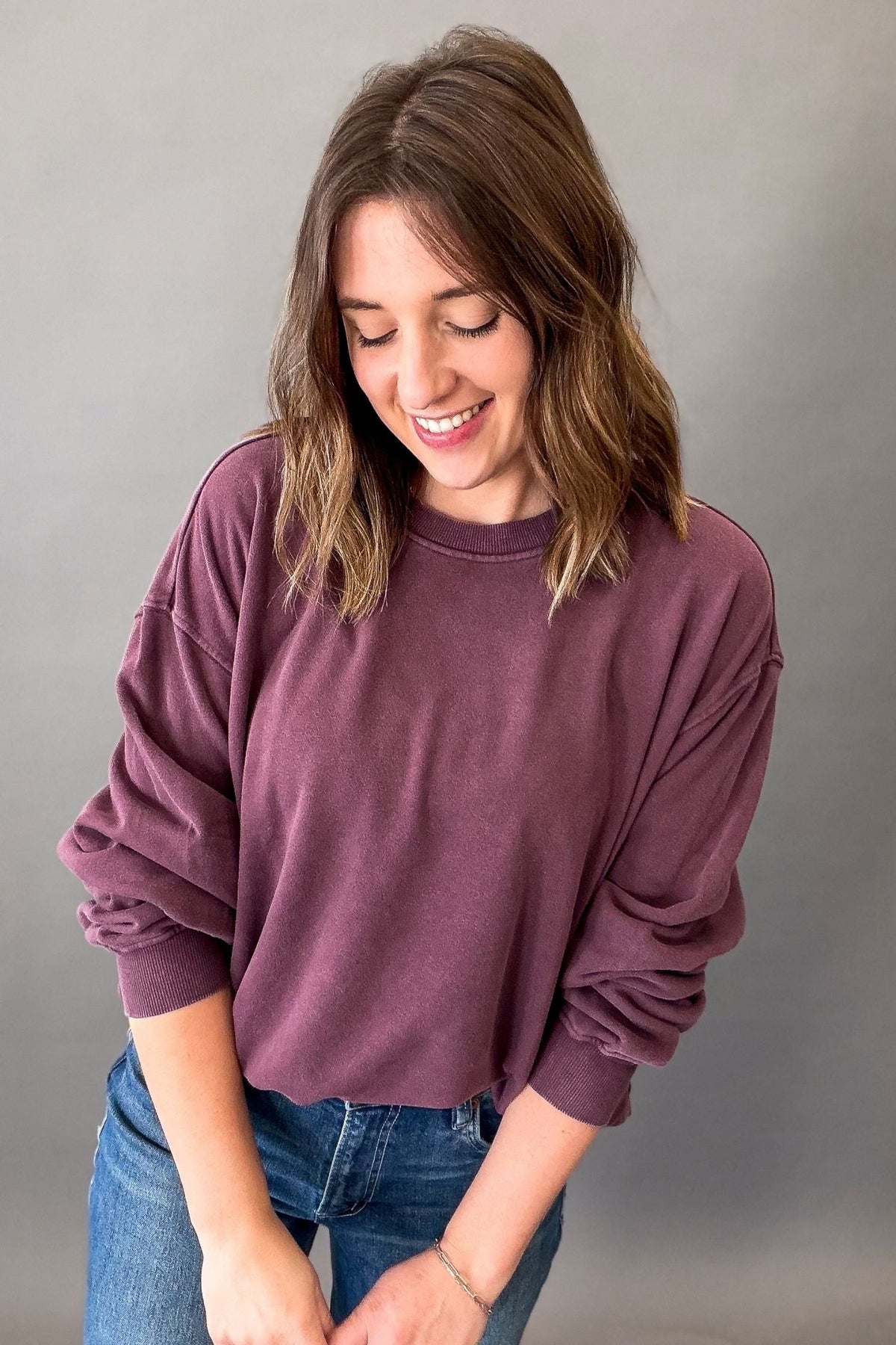 A woman wearing a dusty plum purple long-sleeve crewneck sweatshirt and blue jeans, standing against a neutral gray studio background.