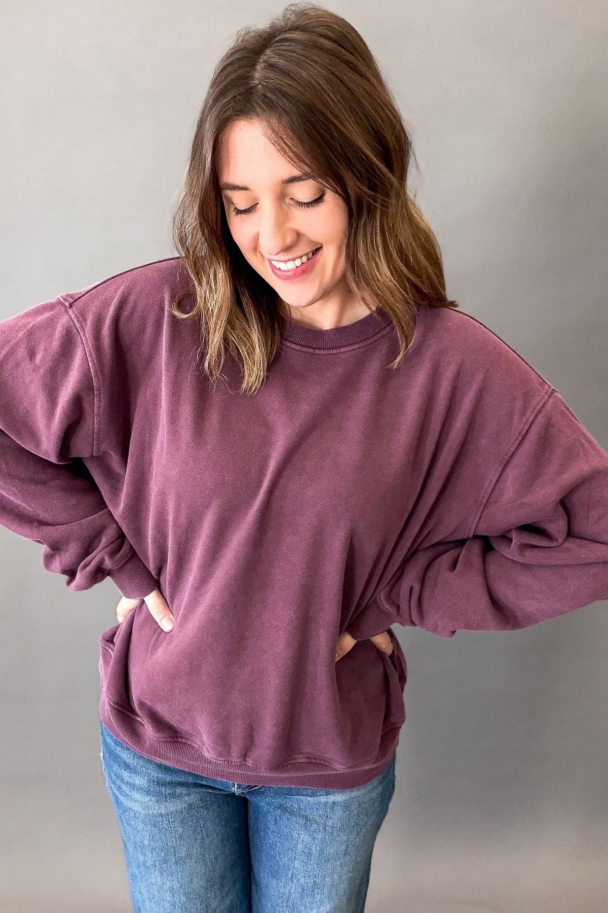 A woman wearing an oversized dusty purple crewneck sweatshirt and blue jeans, standing against a plain gray studio background.