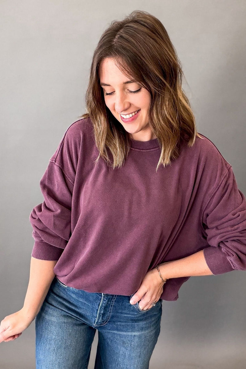 A woman wearing a dusty plum purple long-sleeve crewneck sweatshirt and blue jeans, standing against a neutral gray background.