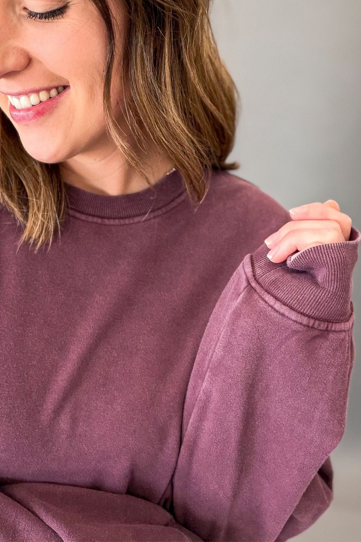 A close-up of a woman wearing a dusty mauve crewneck sweatshirt, showing the ribbed collar and sleeve cuffs, with her hand resting on her shoulder.
