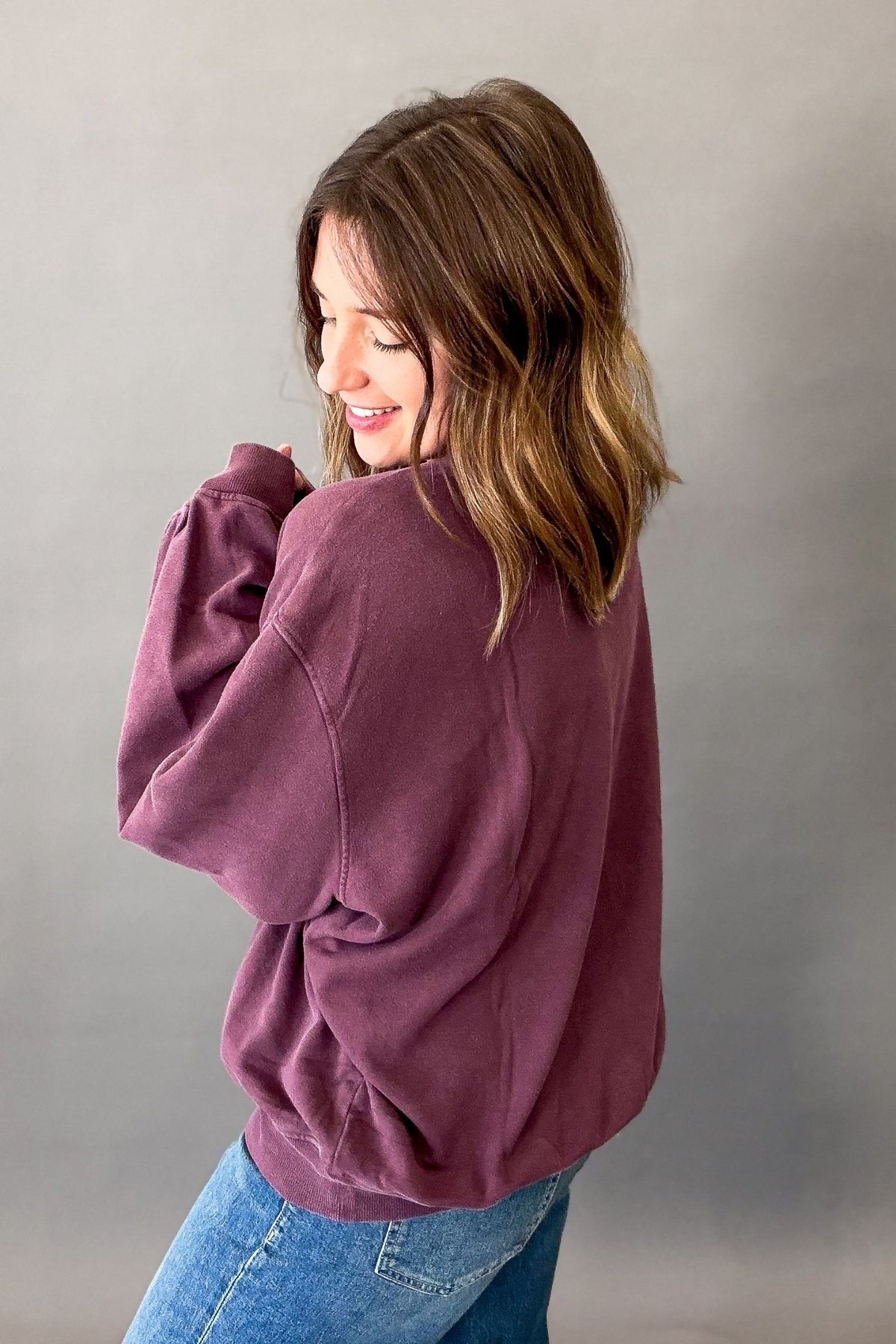 A woman wearing an oversized dusty plum purple sweatshirt and blue jeans, shown from a side-back angle against a neutral gray studio background.