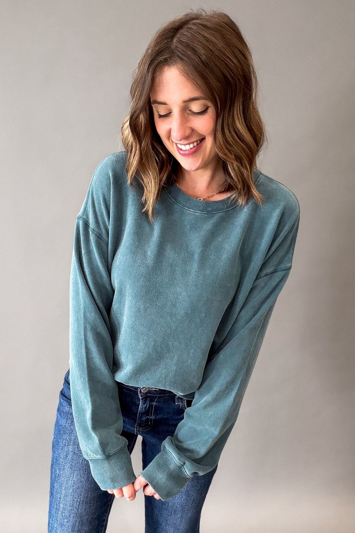A woman wearing a acid wash dark teal long-sleeve crewneck sweatshirt and blue jeans, smiling and looking down against a plain gray studio background.