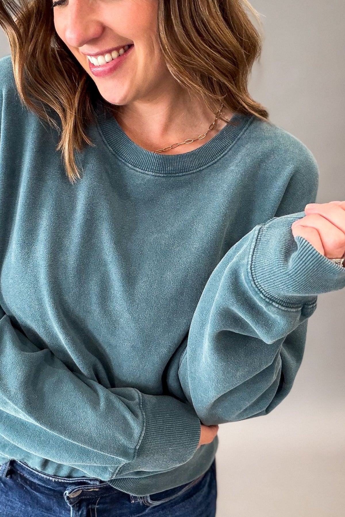 A woman wearing a dark teal acid wash crewneck sweatshirt and blue jeans, shown from the chest up against a plain light gray background.