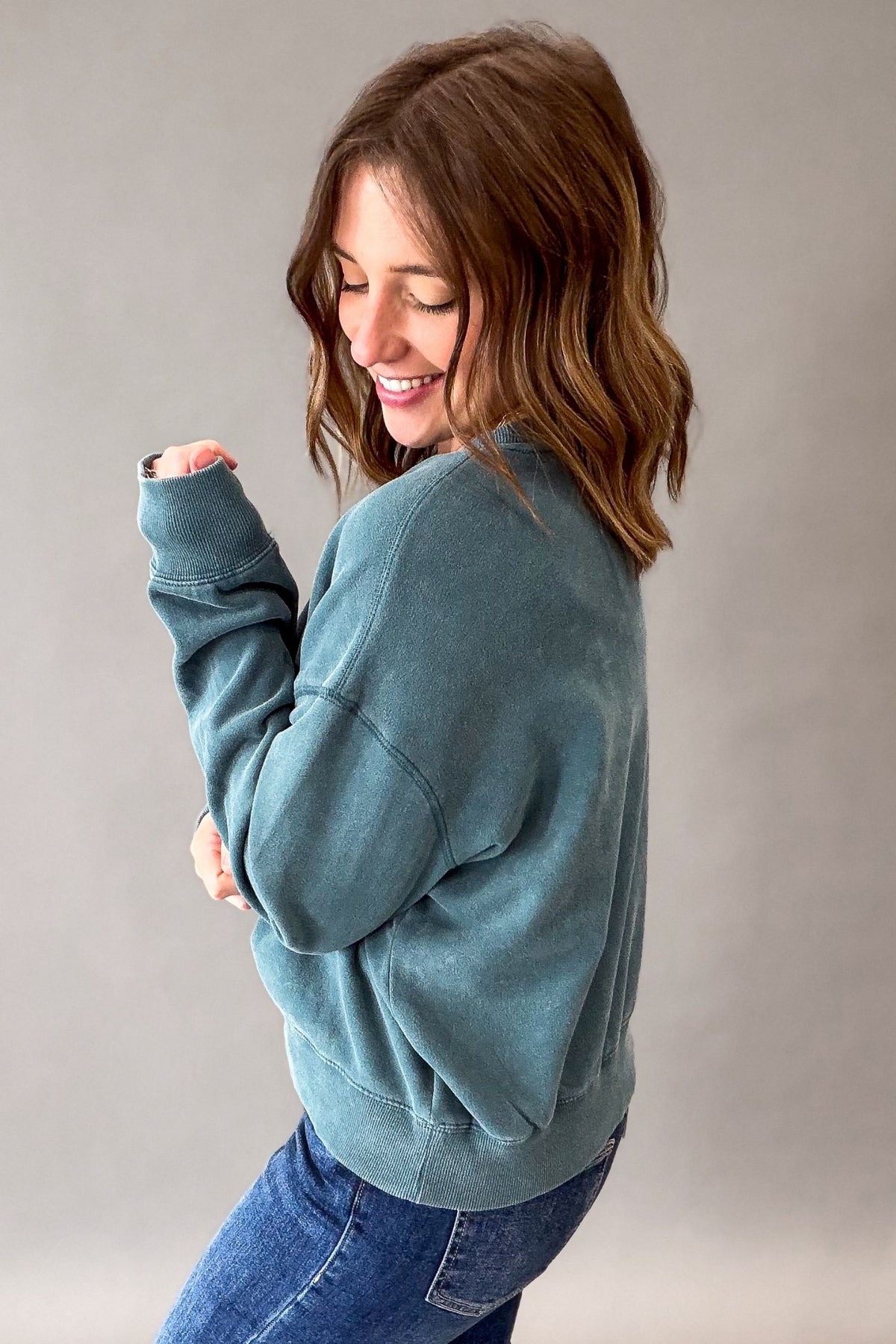 A woman wearing a dark teal acid wash crewneck sweatshirt and blue jeans, shown from a side-back angle against a neutral gray studio background.