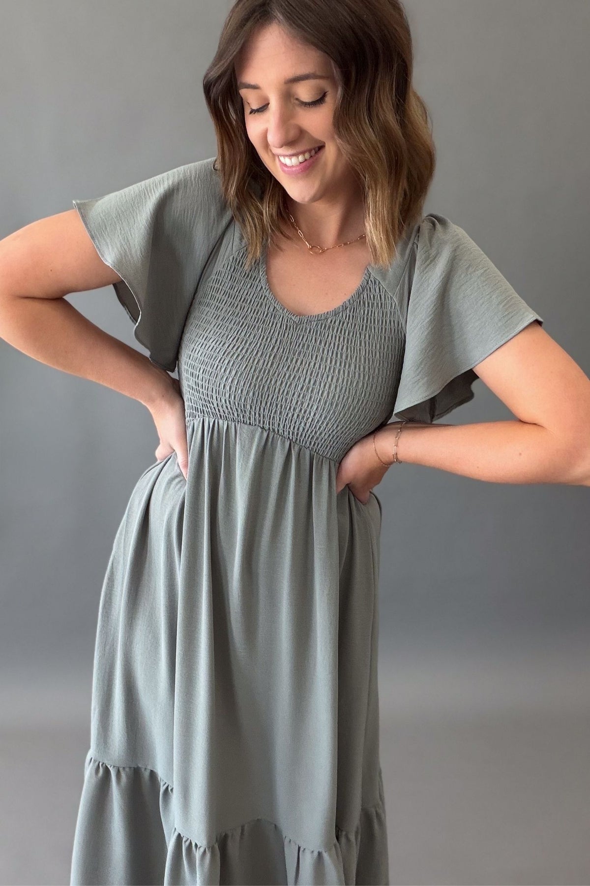 Woman wearing a dusty sage midi dress with a smocked bust and a tiered bottom with flutter sleeves