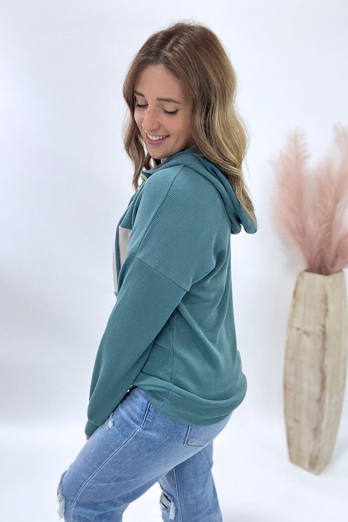 Woman wearing a teal hoodie and blue jeans standing against a white background with a decorative vase.