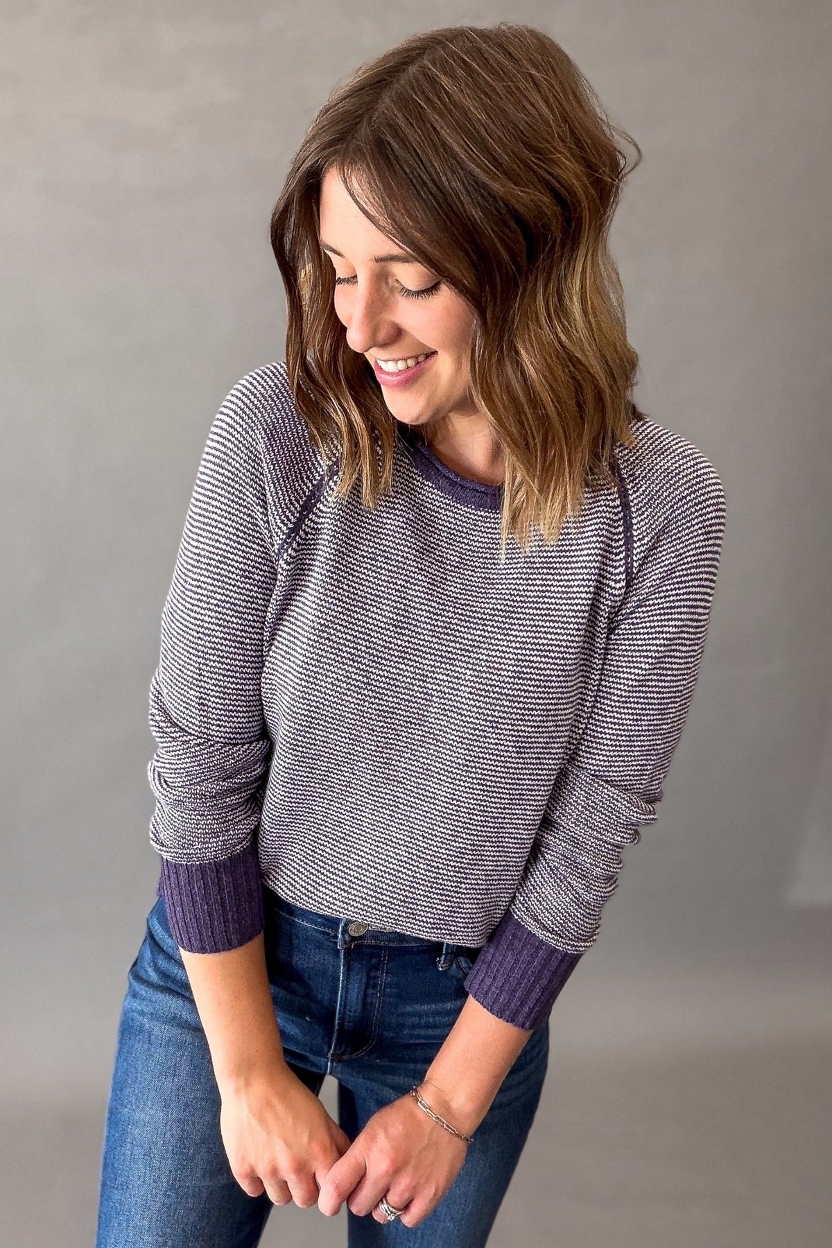 A woman wearing a purple striped long-sleeve crewneck sweater with solid purple ribbed cuffs and collar, paired with blue jeans, standing against a neutral gray studio background.