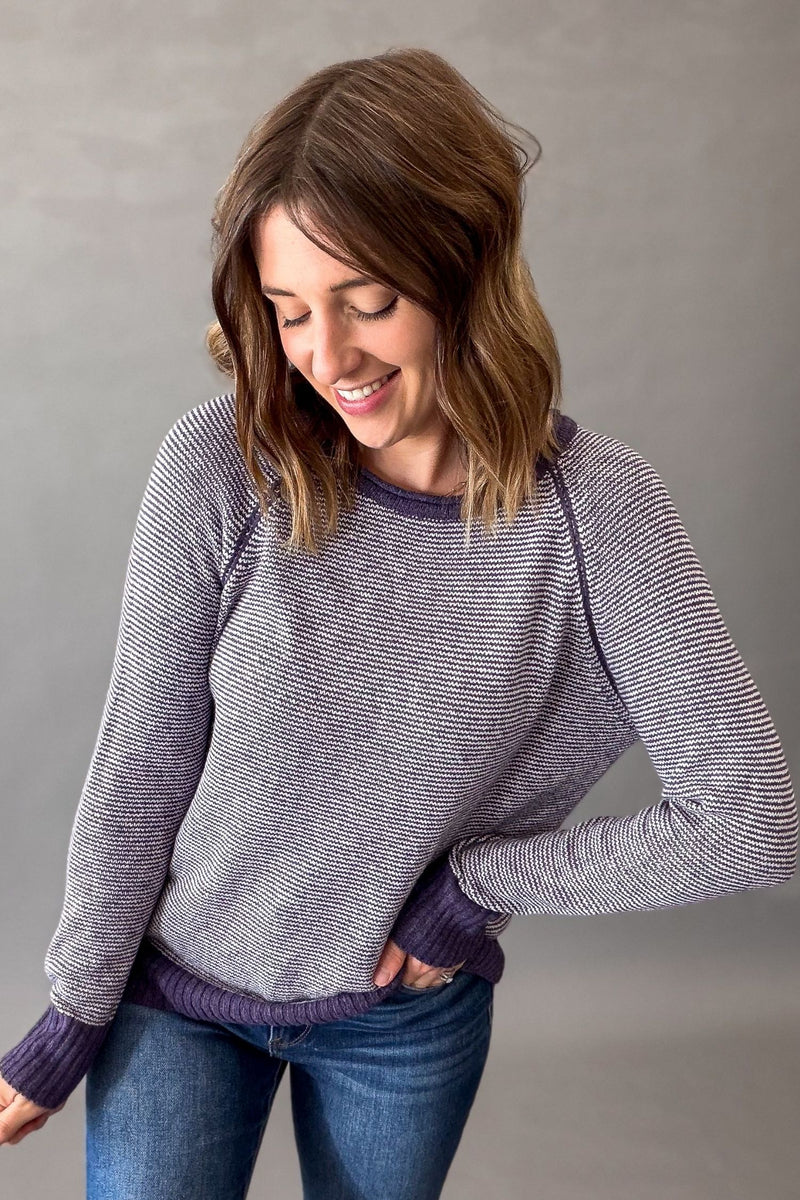 A woman wearing a purple striped long-sleeve knit sweater with solid purple ribbed trim, paired with blue jeans, standing against a neutral gray background.