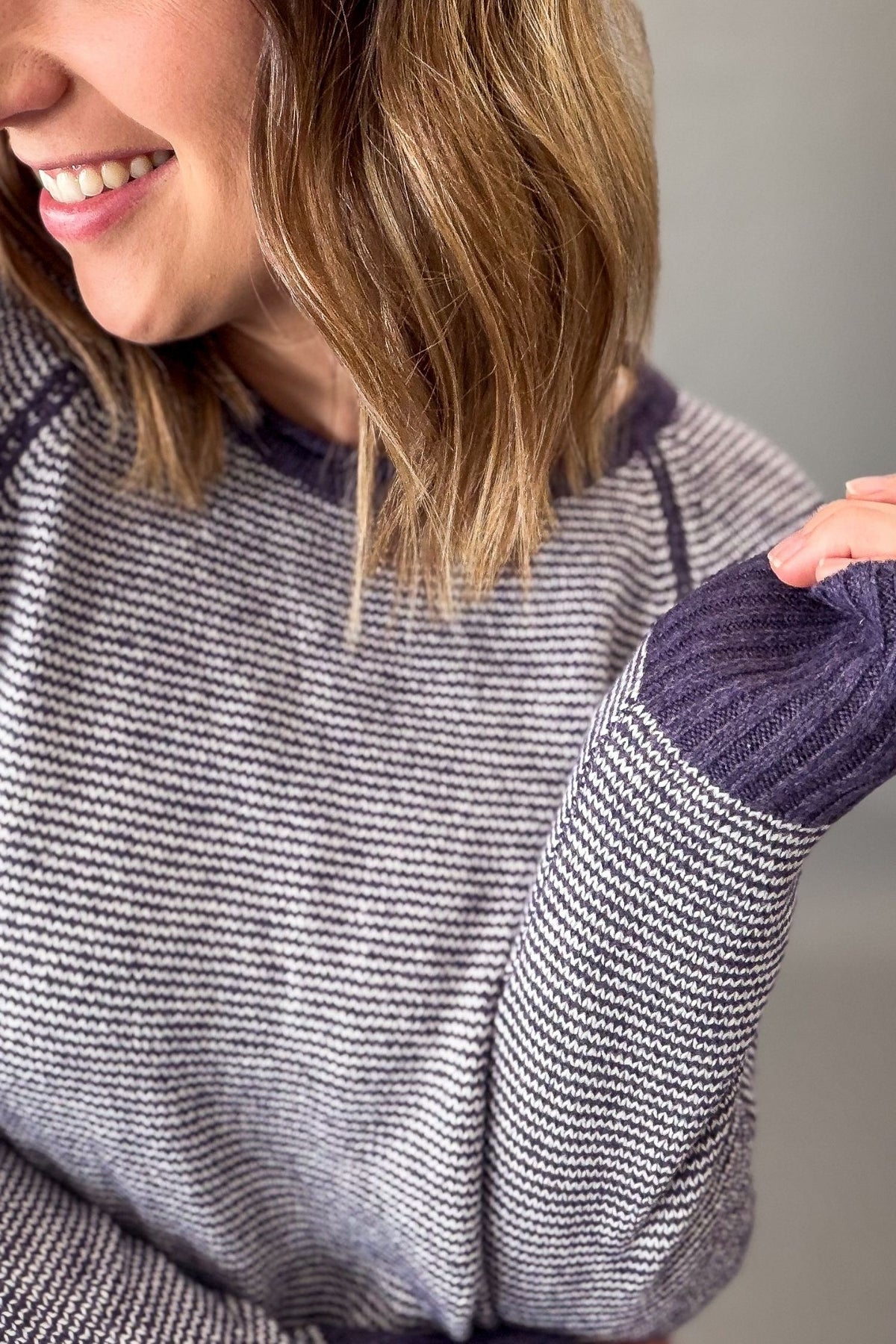 Close-up of a woman wearing a striped knit sweater with contrasting dark purple ribbed cuffs and collar, smiling while pulling the sleeve to show texture.