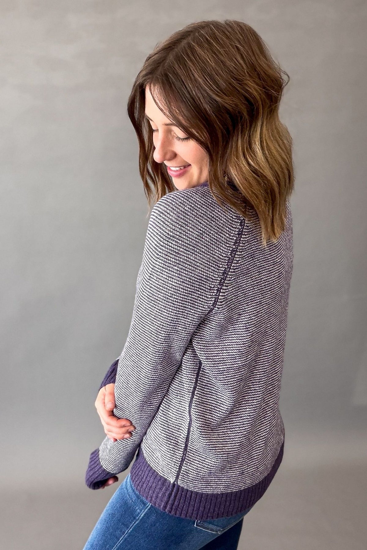 A woman wearing a textured purple knit sweater with blue jeans, photographed from the back against a neutral gray studio background.