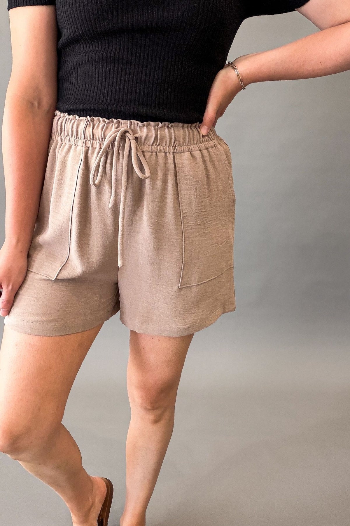 Lightweight taupe women's shorts with a stretchy elastic drawstring waistband, front pockets, and a relaxed fit styled with a black ribbed top.