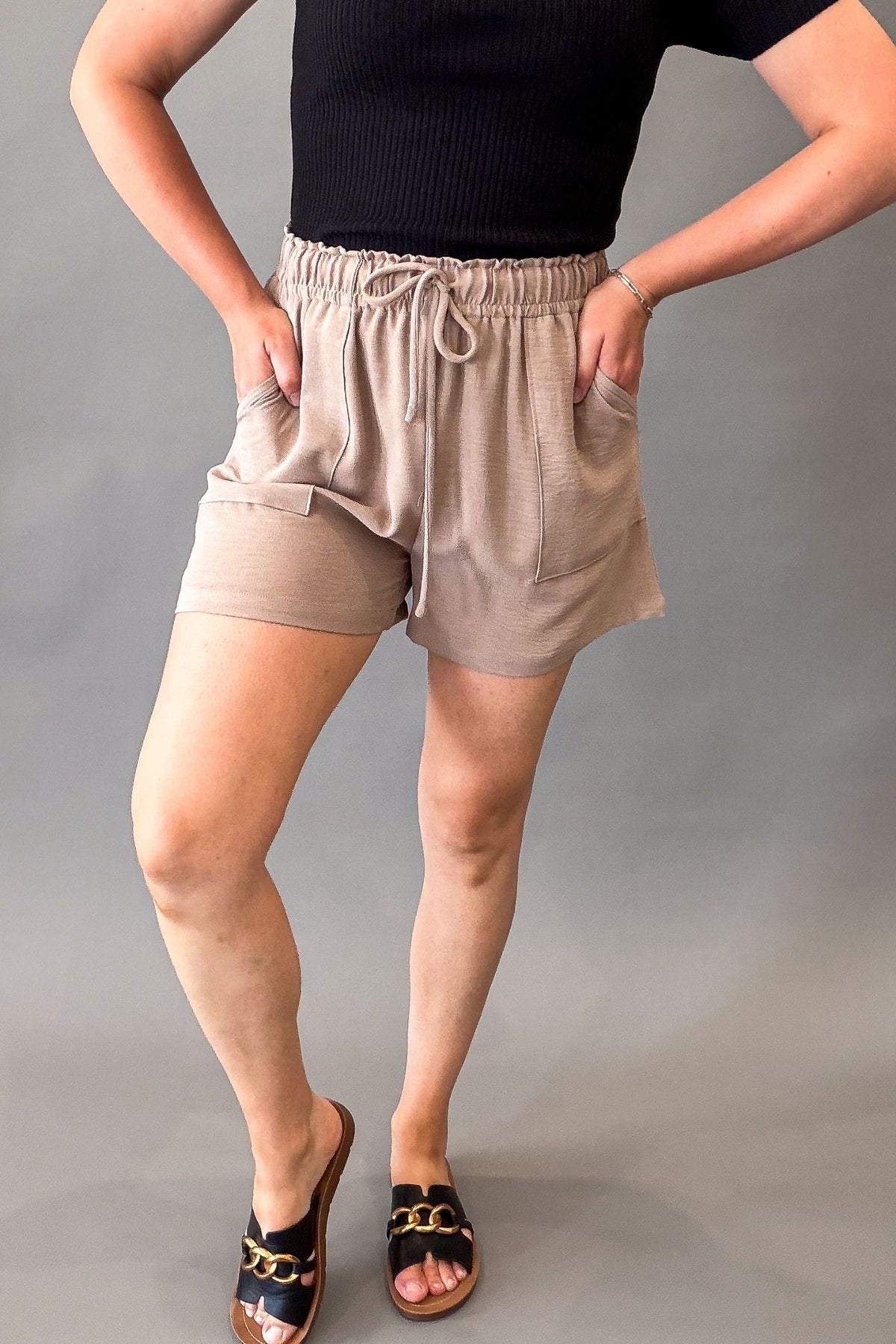 Lightweight taupe women's shorts with a stretchy elastic drawstring waistband, front pockets, and a relaxed fit styled with a black ribbed top.