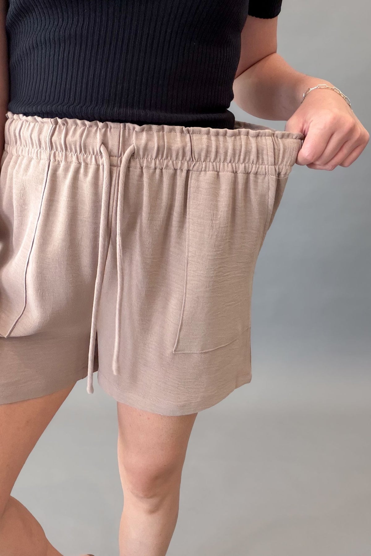 Lightweight taupe women's shorts with a stretchy elastic drawstring waistband, front pockets, and a relaxed fit styled with a black ribbed top showing stretch in the waistband.