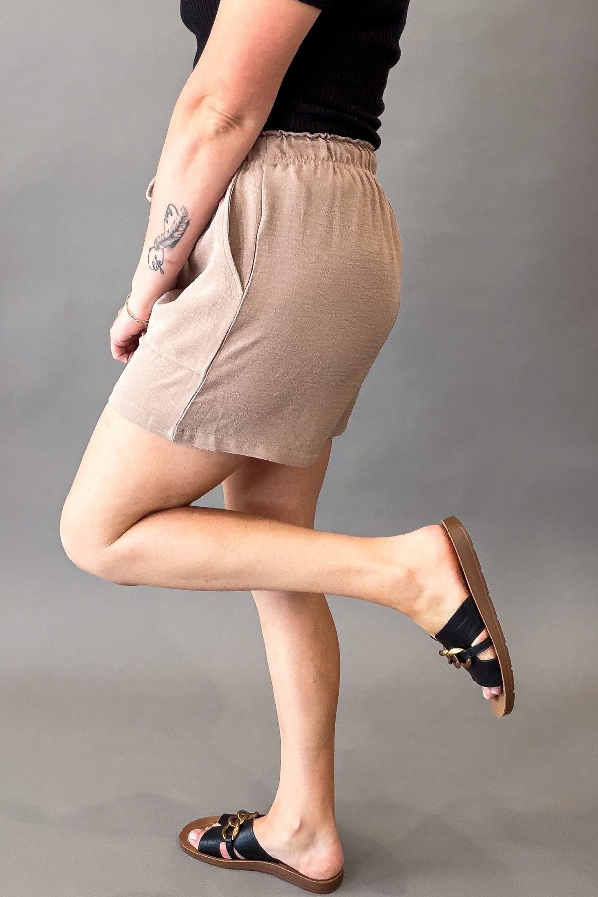 Lightweight taupe women's shorts with a stretchy elastic drawstring waistband, front pockets, and a relaxed fit styled with a black ribbed top showing the side pocket and back details