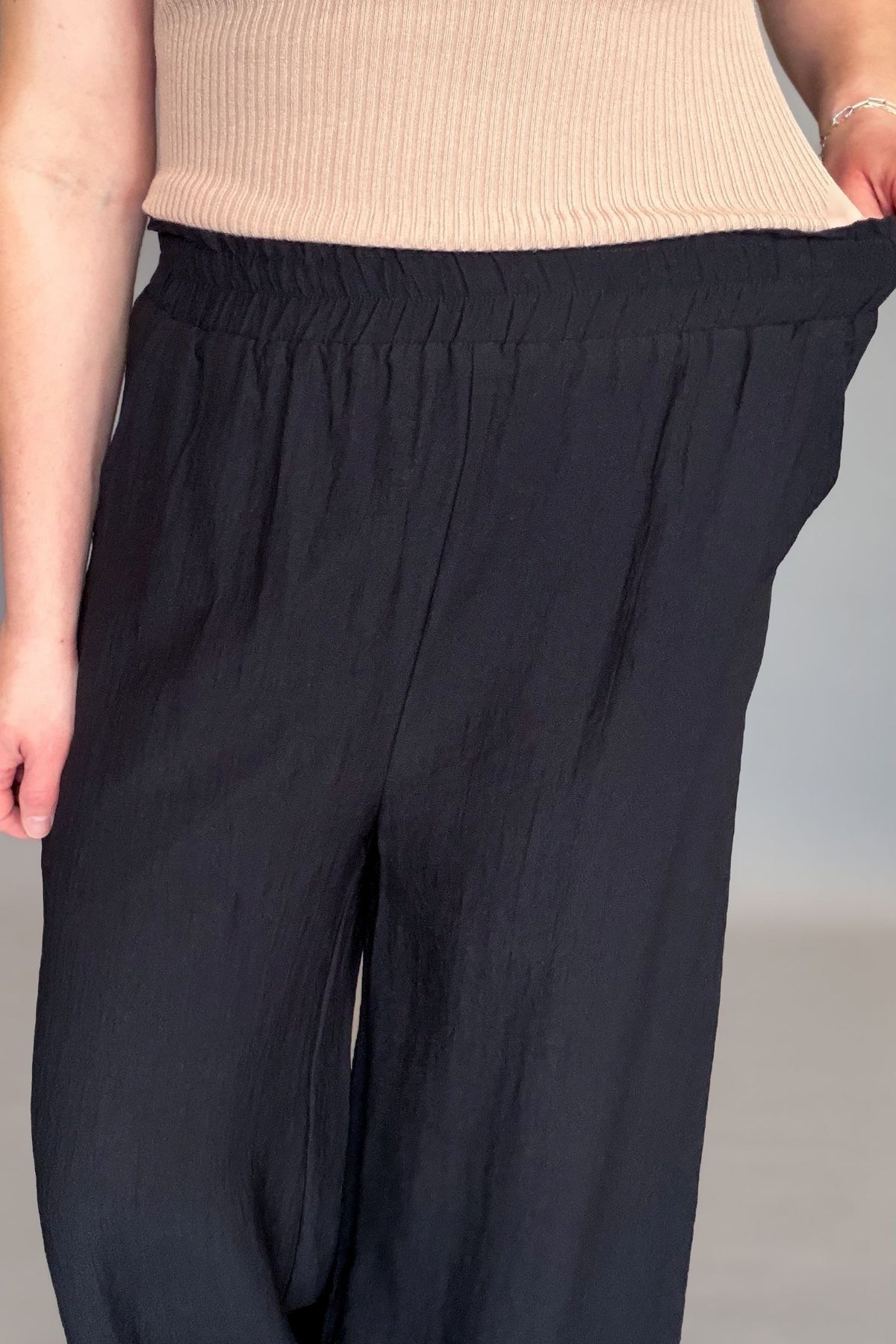 Person wearing black pants and a beige top on a neutral background showing stretch in waistband