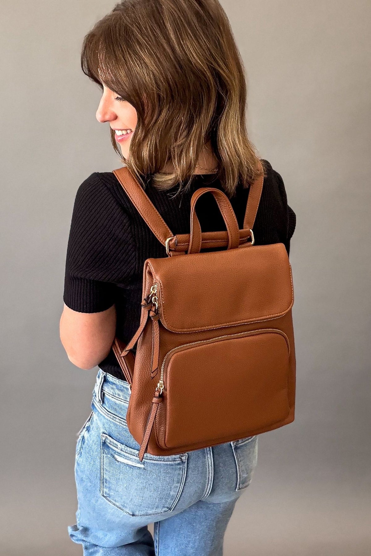 Woman wearing a Brown vegan leather convertible backpack with front and back zip pockets, flap closure, gold hardware, and adjustable straps worn as a stylish everyday backpack.