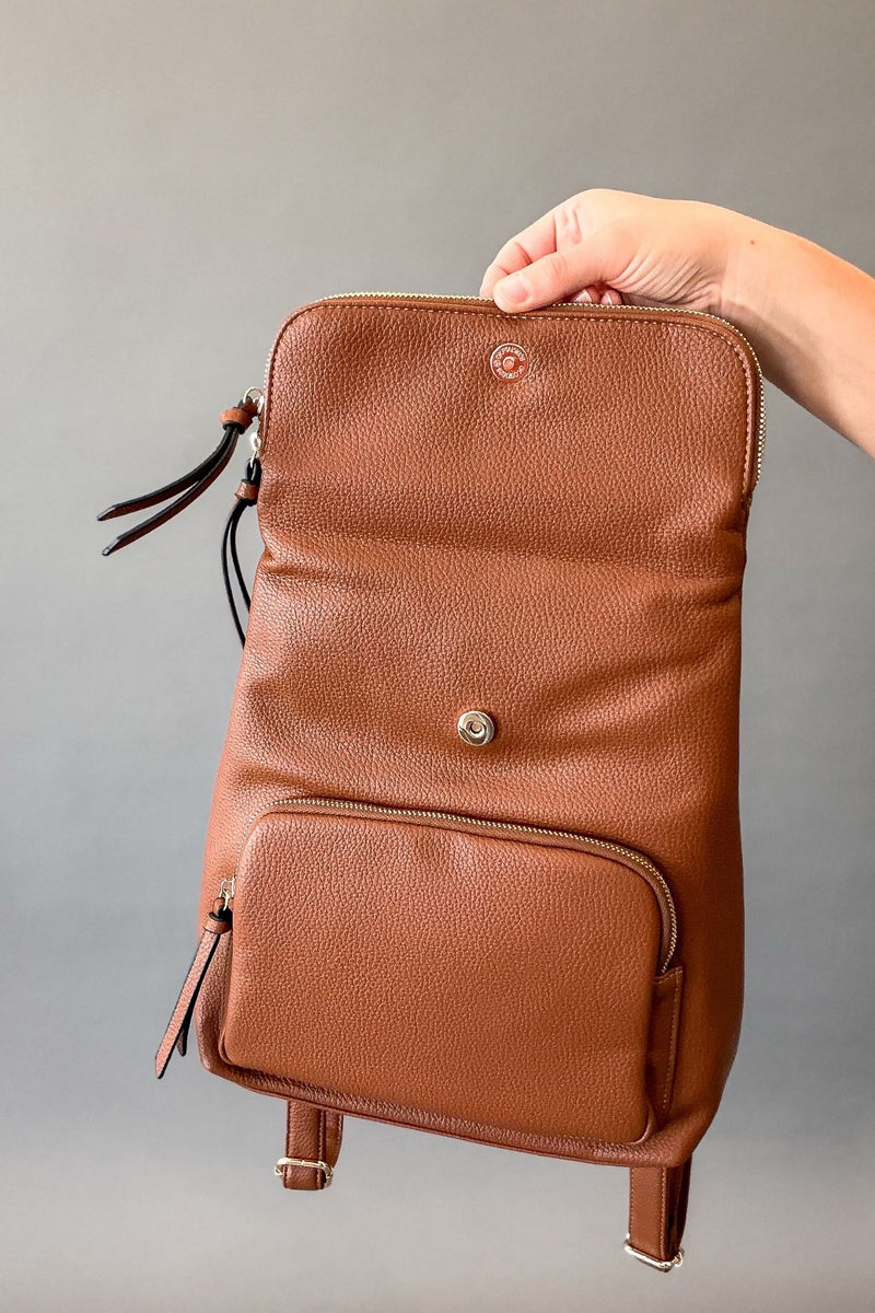 Woman holding a brown backpack open with gold hardware showing the snap button closure, zip top and front zip pocket