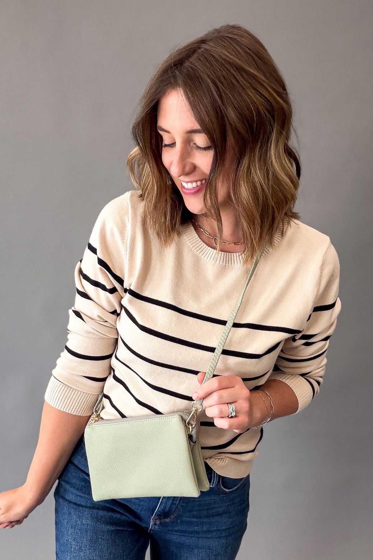 A woman wearing a cream and black striped crewneck sweater, blue jeans, and a light sage green crossbody clutch bag, posing against a solid gray studio background.