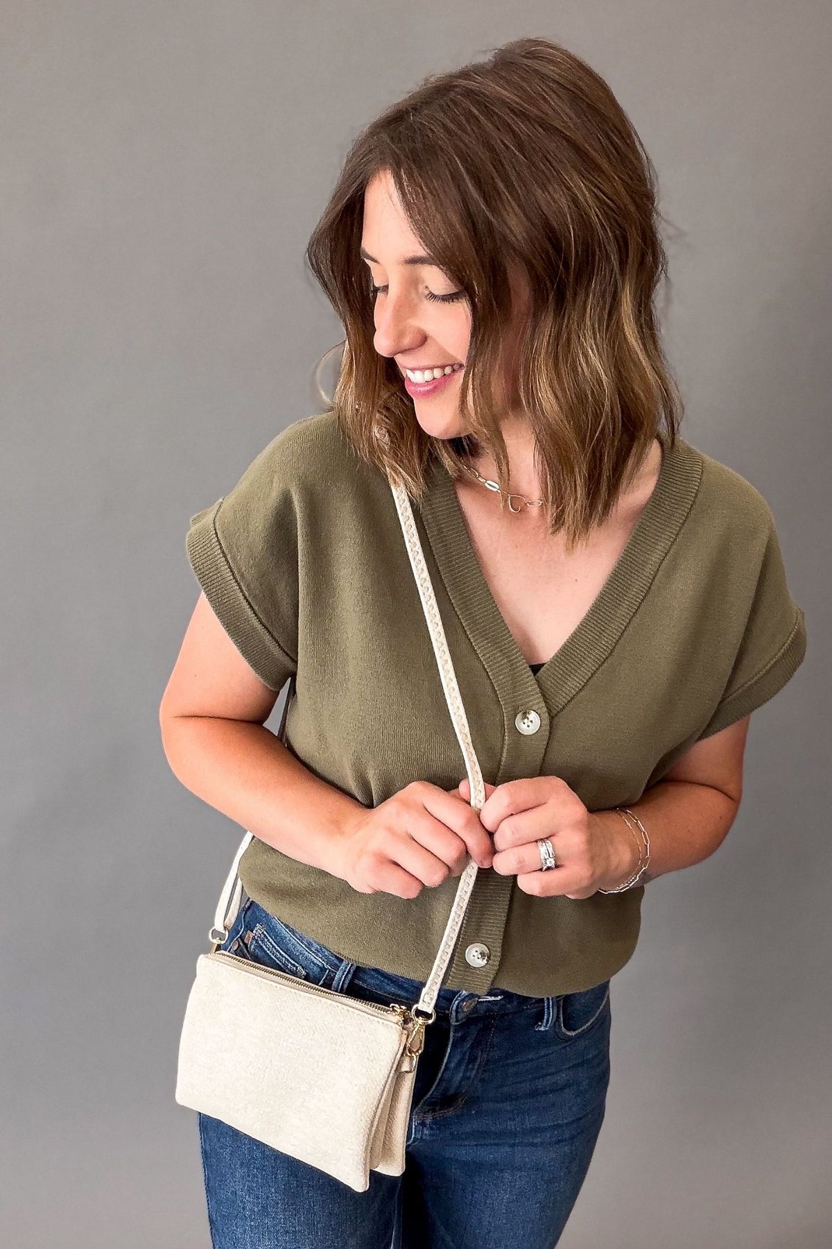 A woman wearing an olive green short-sleeve button-up top and blue jeans, carrying a small cream-colored crossbody bag against a solid gray studio background.