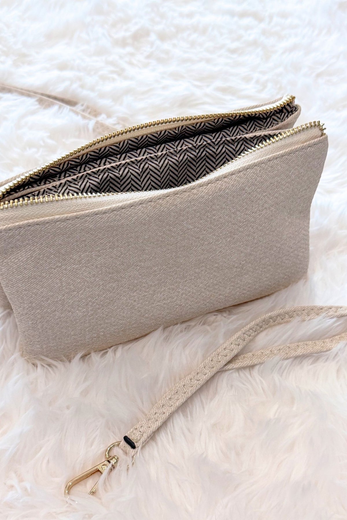 A light beige textured zippered pouch with a matching wrist strap and gold-tone hardware, shown open on a white faux-fur surface.