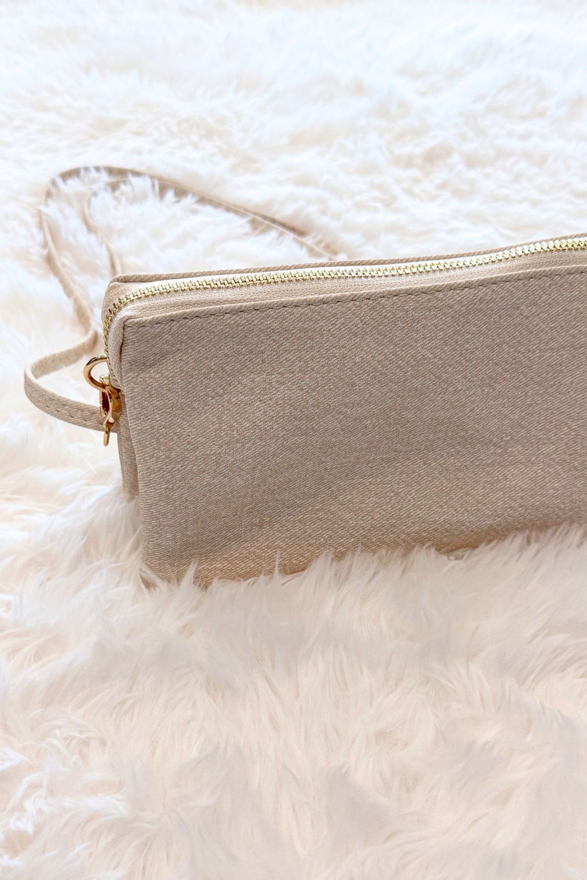 A light beige textured zippered crosbody with a matching wrist strap and gold-tone hardware, resting on a white faux fur surface.