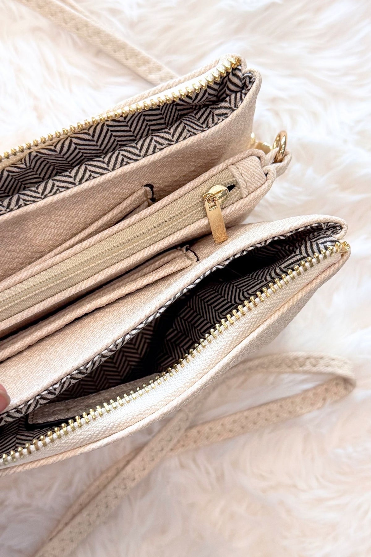 Close-up of a beige textured crossbody or wallet-style handbag with gold-tone zippers and a black-and-beige patterned interior lining, shown on a soft white faux-fur surface.