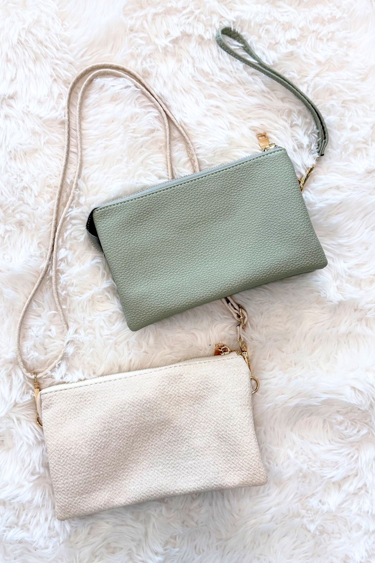 Two small pebbled leather crossbody pouches with wrist straps, one in sage green and one in cream, arranged on a white faux fur background.