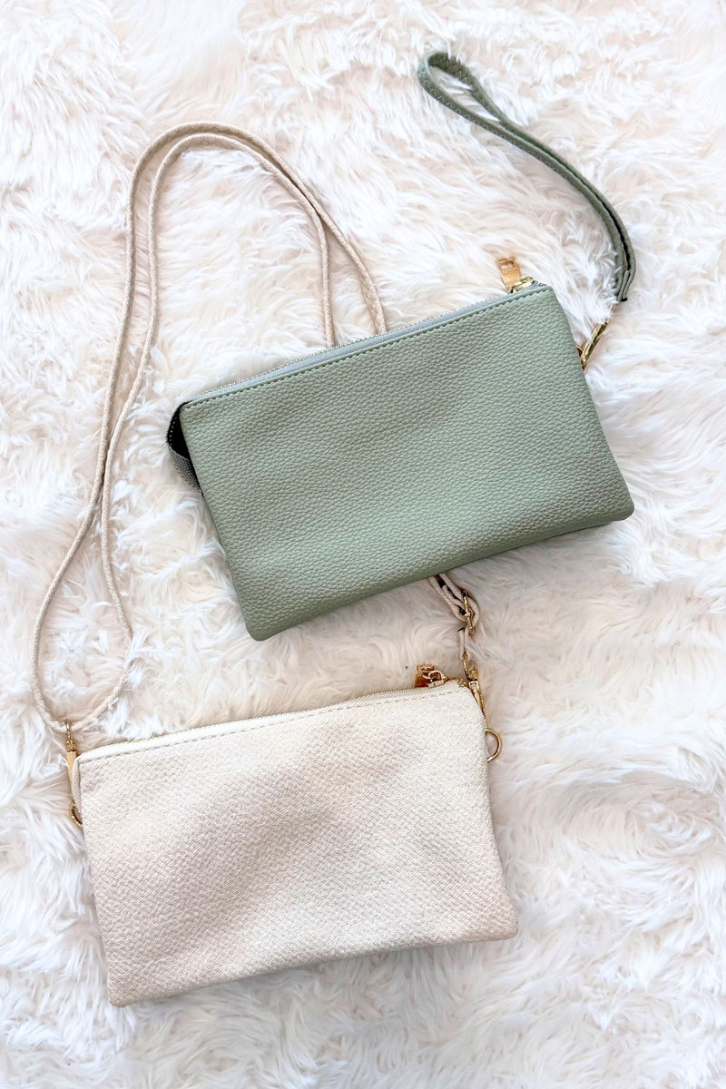 Two small pebbled leather crossbody pouches with wrist straps, one in sage green and one in cream, arranged on a white faux fur background.