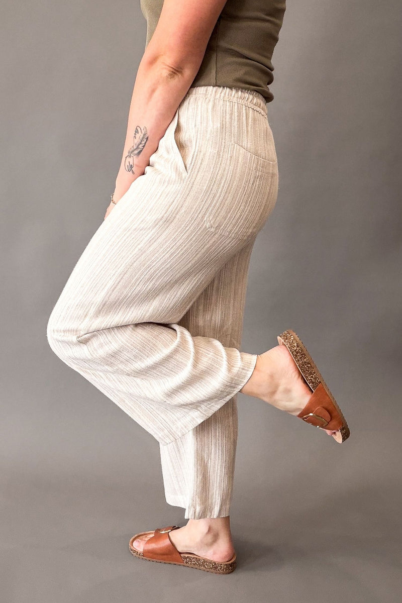 A person wearing loose, light beige ribbed pants with a side pocket, paired with brown leather slide sandals, shown from the waist down against a plain gray studio background.