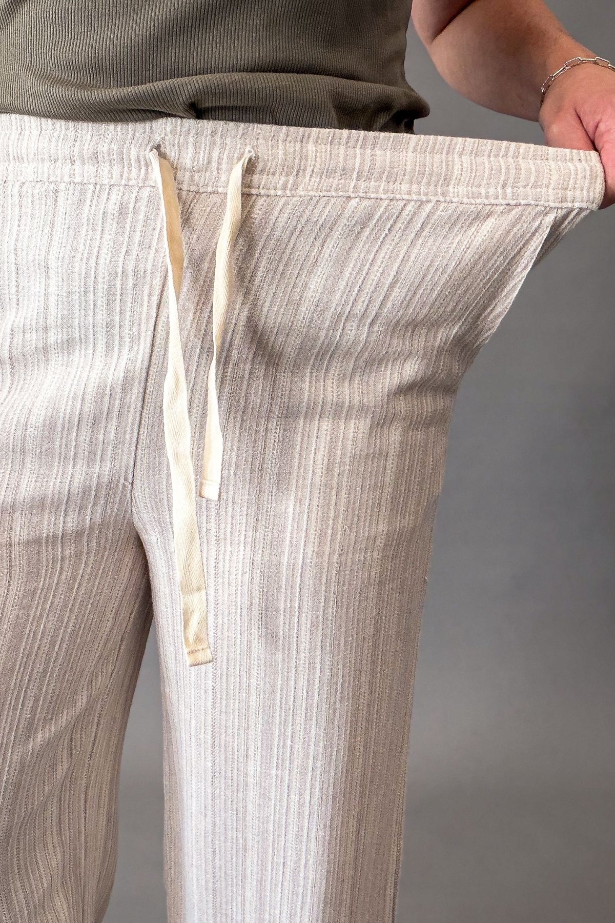 Close-up of a person wearing light beige vertically striped lounge pants, pulling the fabric outward to demonstrate stretch. The pants have a cream-colored drawstring and are shown against a neutral gray background.