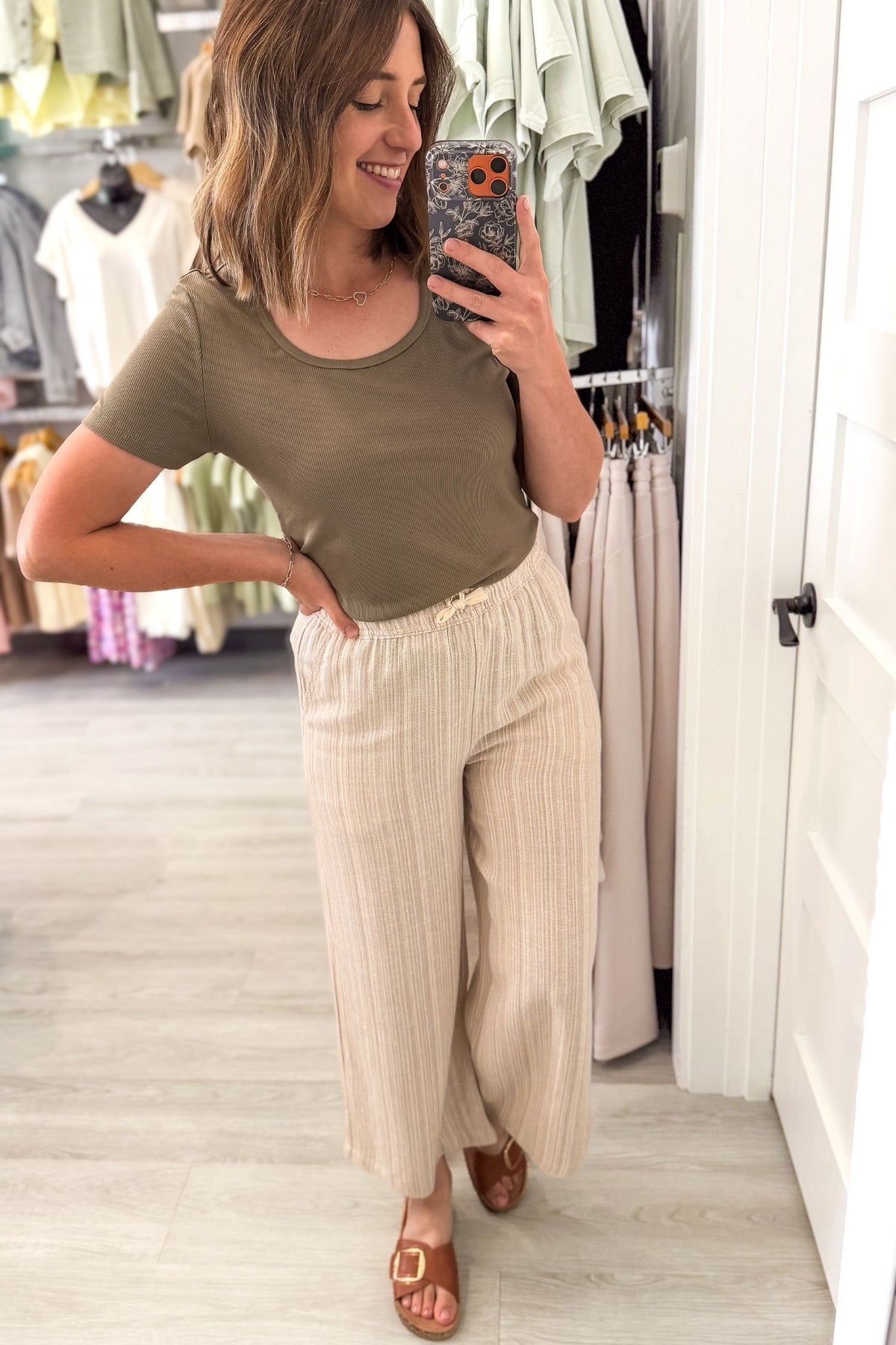 A woman takes a mirror selfie in a clothing boutique, wearing a short-sleeve olive green ribbed knit top and cream-colored wide leg linen blend pants.
