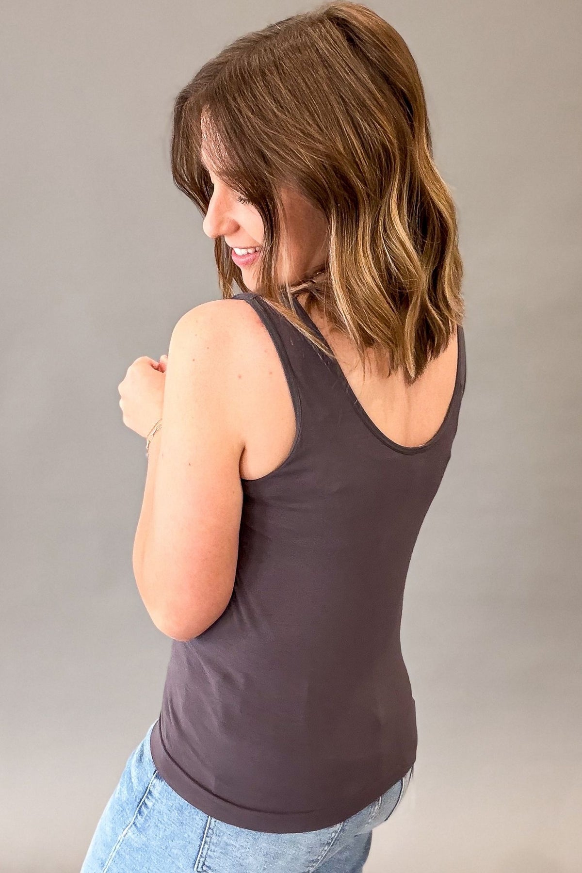 A woman with shoulder-length brown hair is shown from the back wearing a dark charcoal gray ribbed tank top and light blue jeans, set against a plain gray studio background.