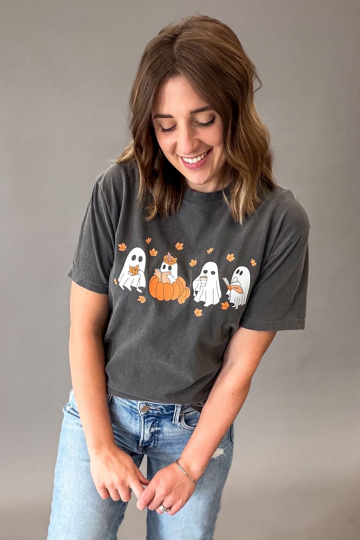 A woman wearing a dark gray graphic t-shirt with a cartoon ghost and pumpkin design, paired with light blue jeans, posing against a neutral gray studio background.
