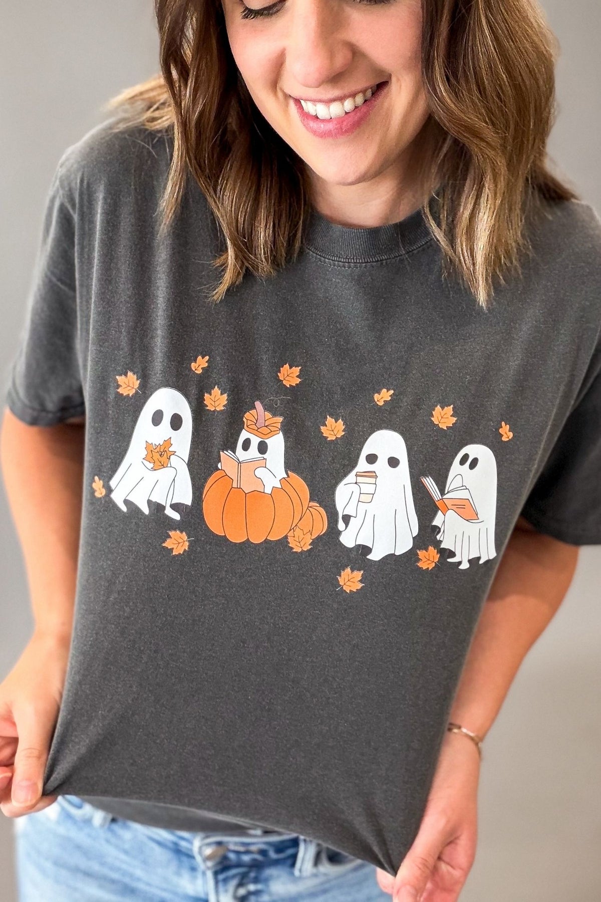 A woman wearing a dark gray graphic t-shirt with a design of ghosts, pumpkins, and orange leaves, paired with blue jeans.