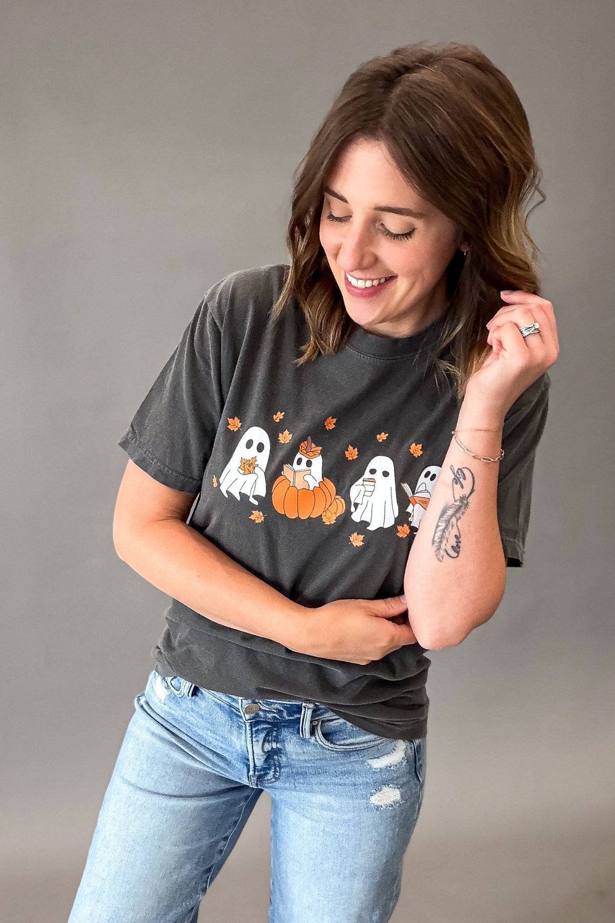 A woman wearing a dark gray graphic t-shirt with a Halloween design of ghosts, pumpkins, and orange leaves, paired with light blue jeans, posing against a solid gray studio background.