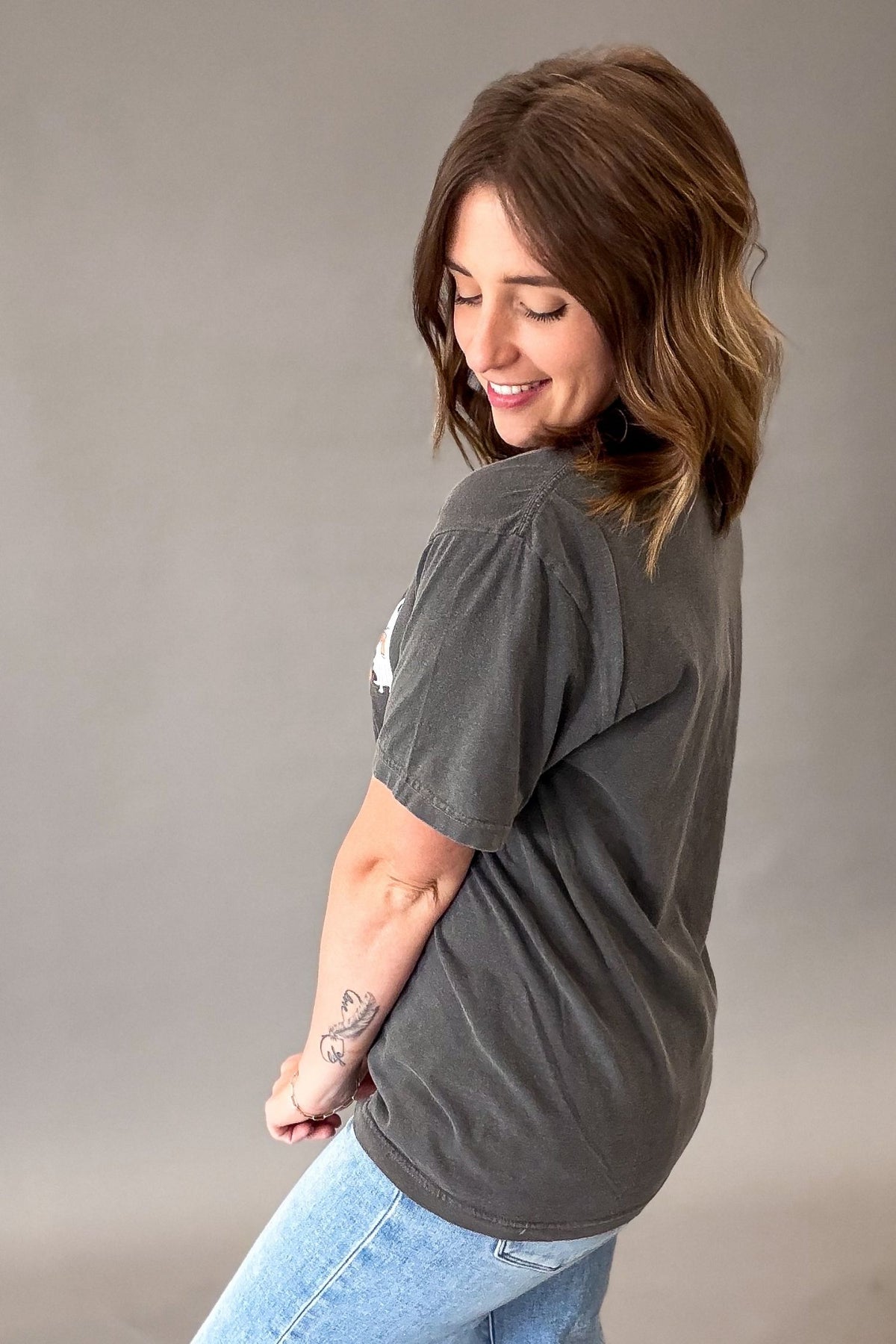 A woman with shoulder-length brown hair is shown from a side-back angle wearing a dark charcoal gray short-sleeve t-shirt and light blue jeans against a solid gray studio background.
