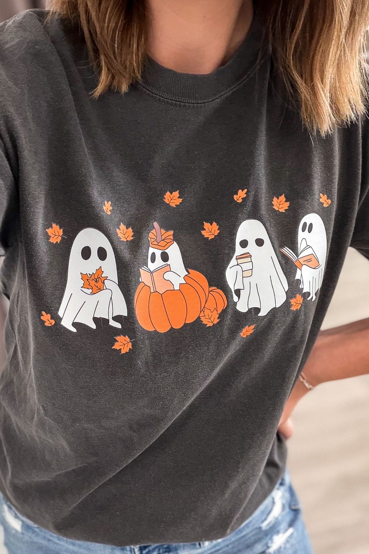 A close-up of a person wearing a dark charcoal gray graphic t-shirt with a Halloween design featuring four white ghosts, some reading, an orange pumpkin, and orange maple leaves.