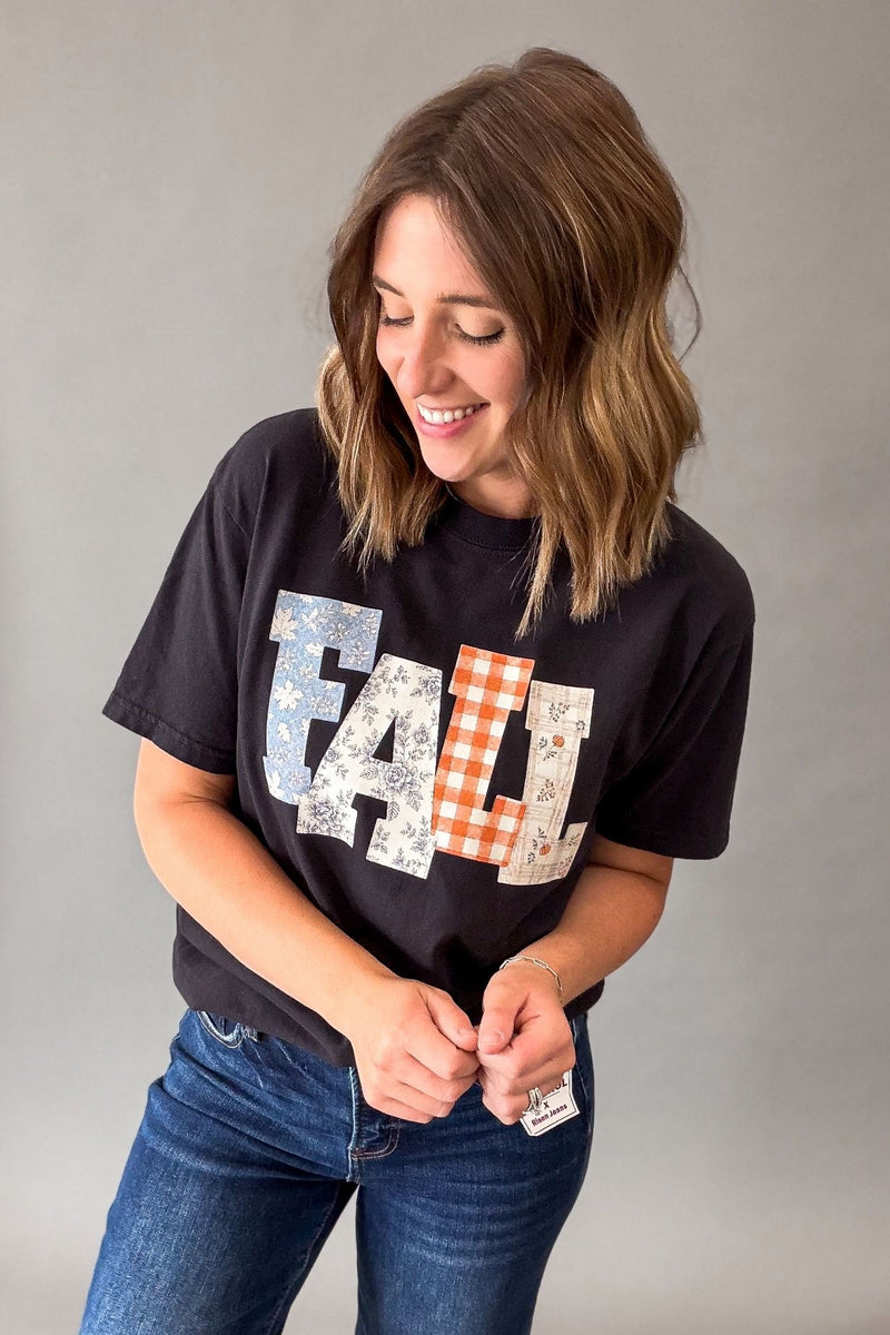 A woman wearing a black graphic t-shirt with a large 'FALL' text design in different patterns, paired with blue jeans, standing against a neutral gray background.