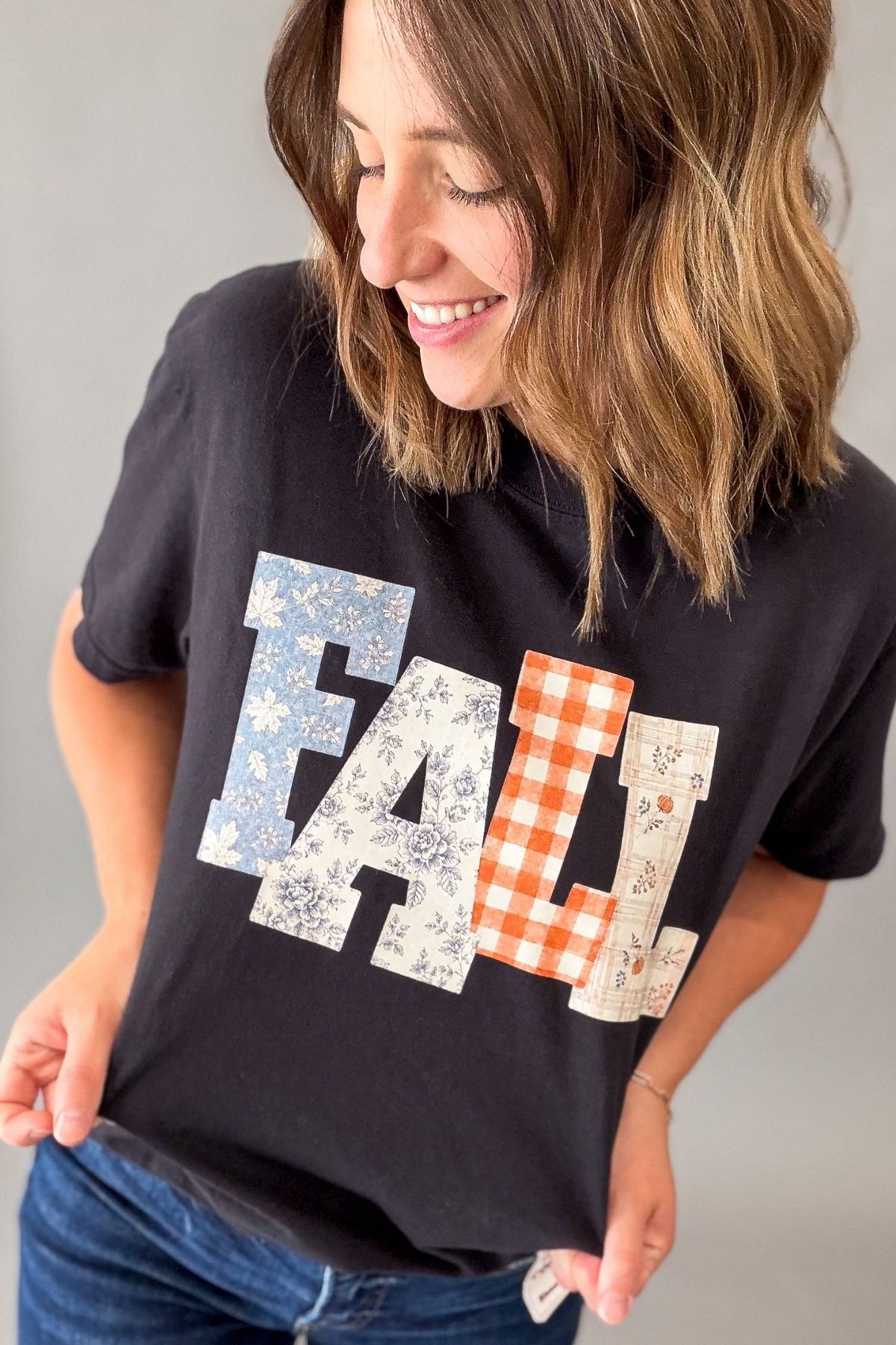 A woman wearing a black graphic t-shirt with the word 'FALL' printed in a patchwork pattern of blue floral, orange-and-white gingham, and beige botanical prints, paired with blue jeans against a neutral gray background.