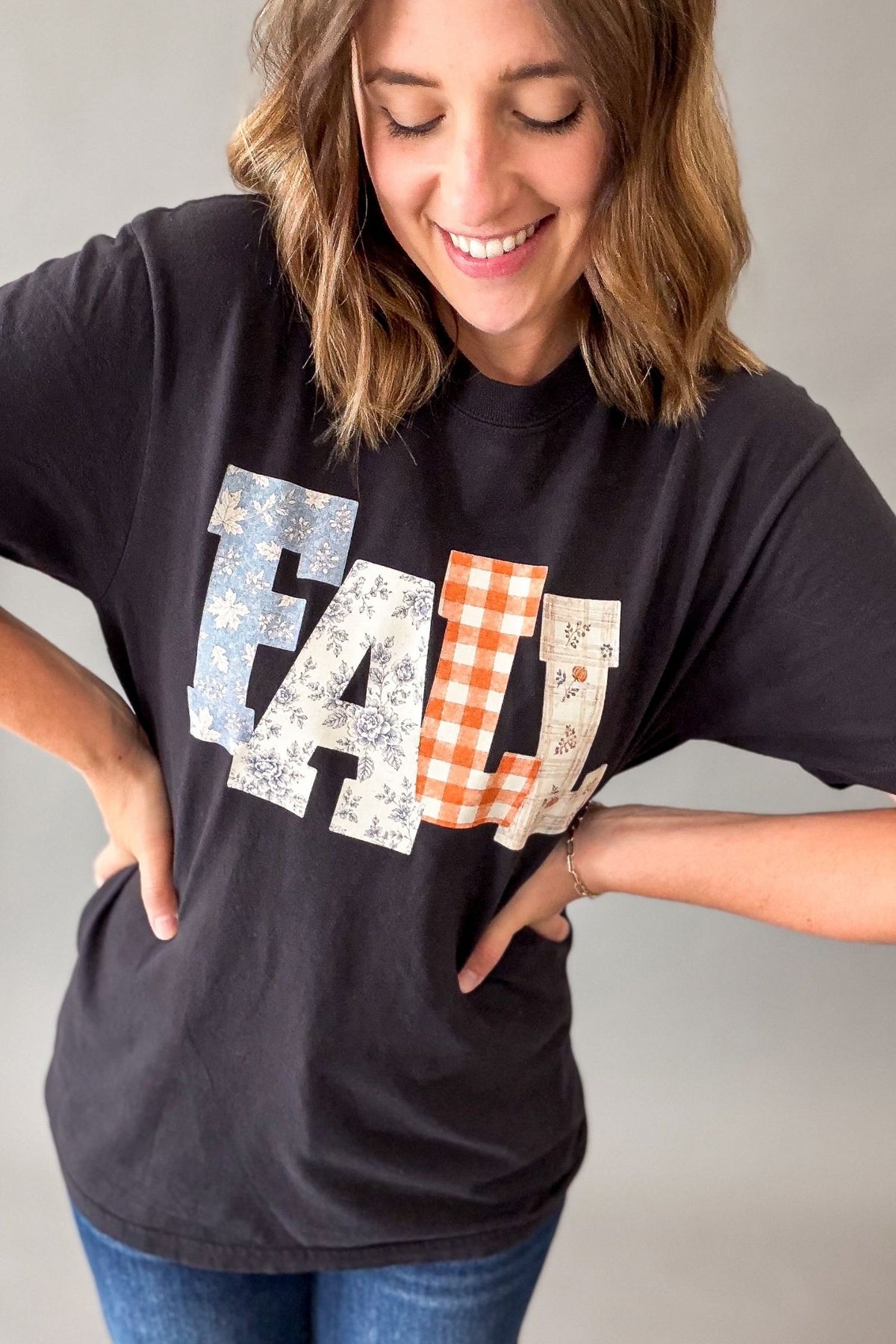 A woman wearing a black graphic t-shirt with the word 'FALL' spelled out in patchwork patterns of blue floral, orange gingham, and cream floral. She is smiling and posing with her hands on her hips against a neutral gray background.