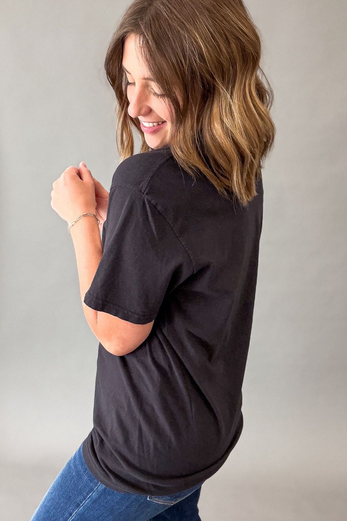 A woman with shoulder-length brown hair is shown from a side-back angle wearing a black short-sleeve crewneck t-shirt and blue jeans against a neutral gray studio background.
