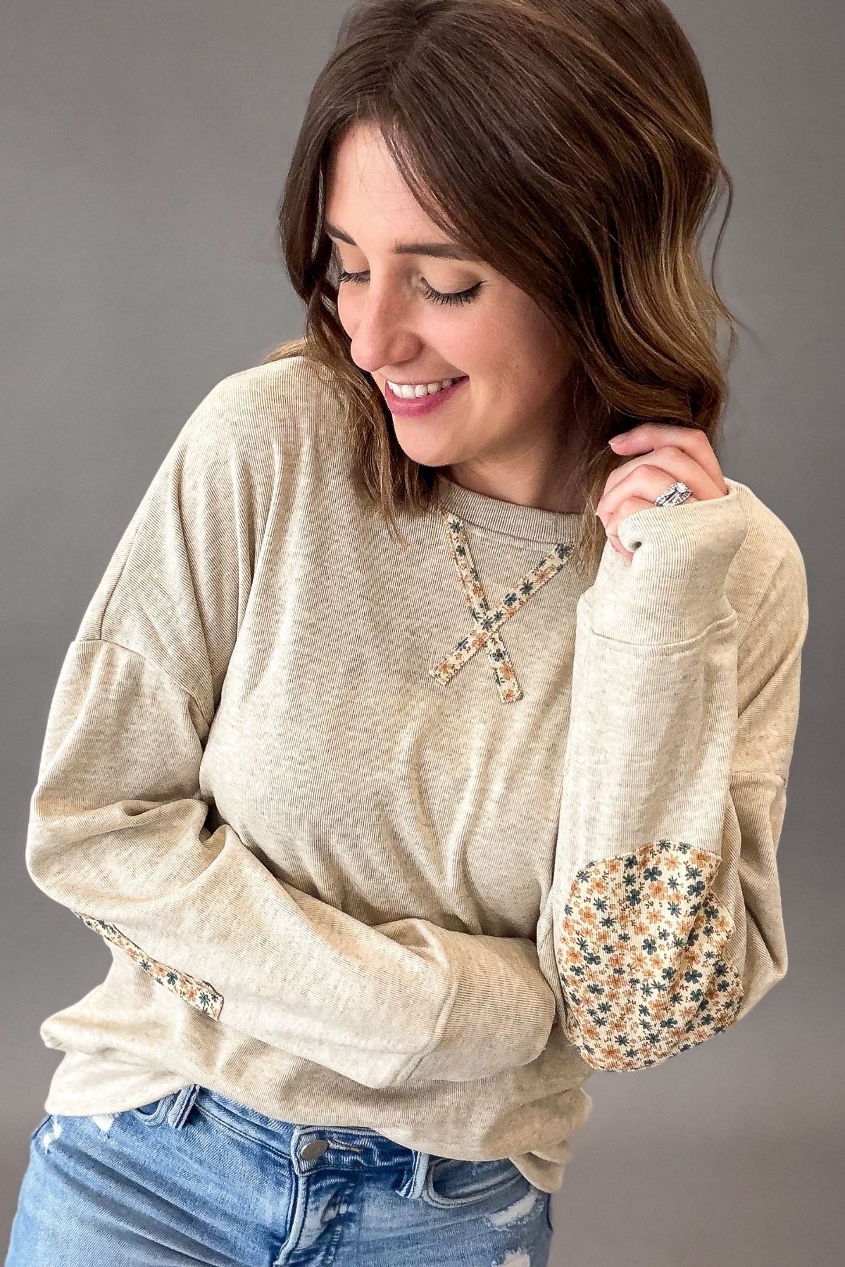A woman wearing a oatmeal long-sleeve top with floral elbow patches and a matching floral cross detail on the chest, paired with blue jeans, against a solid gray studio background.